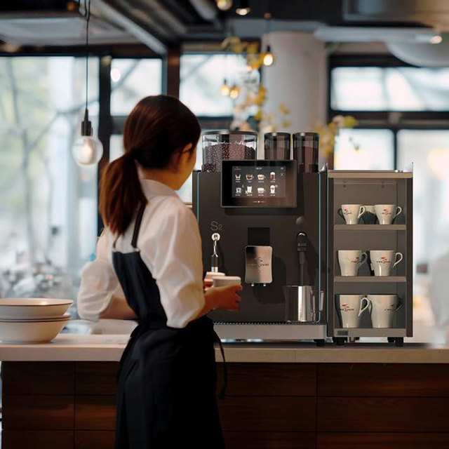 Rex-Royal S2 bean-to-cup coffee machine, designed for cafés, restaurants, and offices. Compact and Swiss-engineered, it features a 7-inch touchscreen, advanced milk system, and automated cleaning. Customisable with payment solutions and cup dispensers, delivering barista-quality coffee efficiently in high-demand environments.