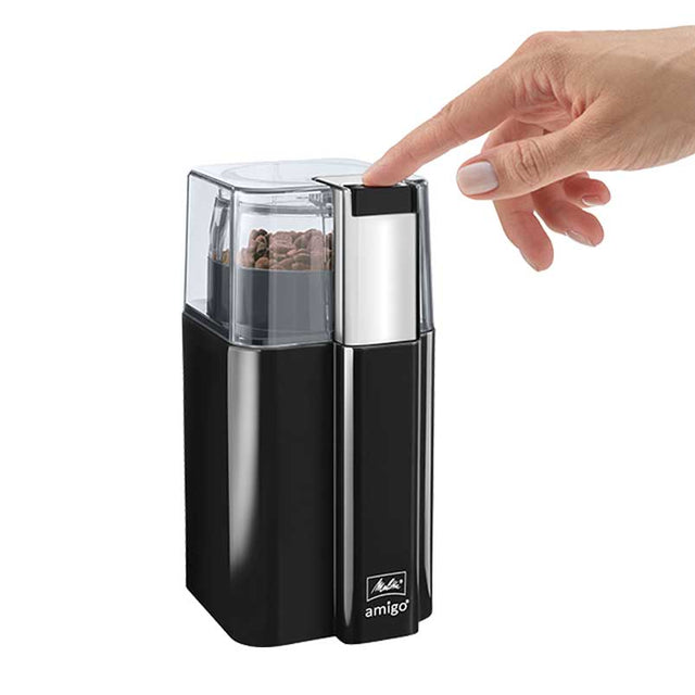 A hand pressing the button on the Melitta AMIGO Electric Blade Coffee Grinder, showing coffee beans inside the transparent lid and the grinder's sleek black and chrome design.