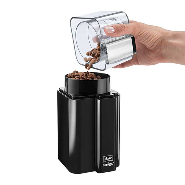 A hand pouring coffee beans from a transparent lid into the Melitta AMIGO Electric Blade Coffee Grinder, showcasing the grinder's compact black design and user-friendly bean container lid.